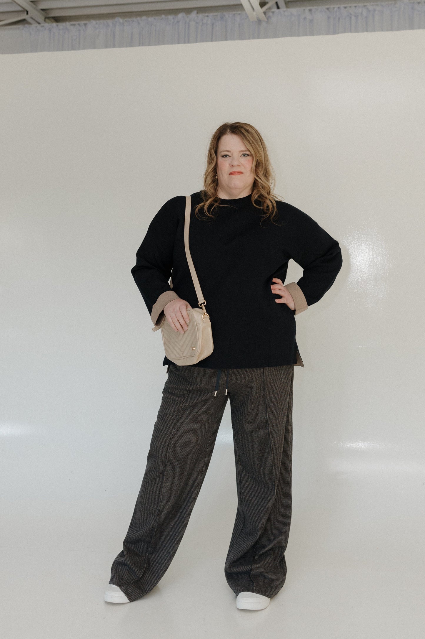 Woman wearing a black sweater and gray pants standing against a white wall.