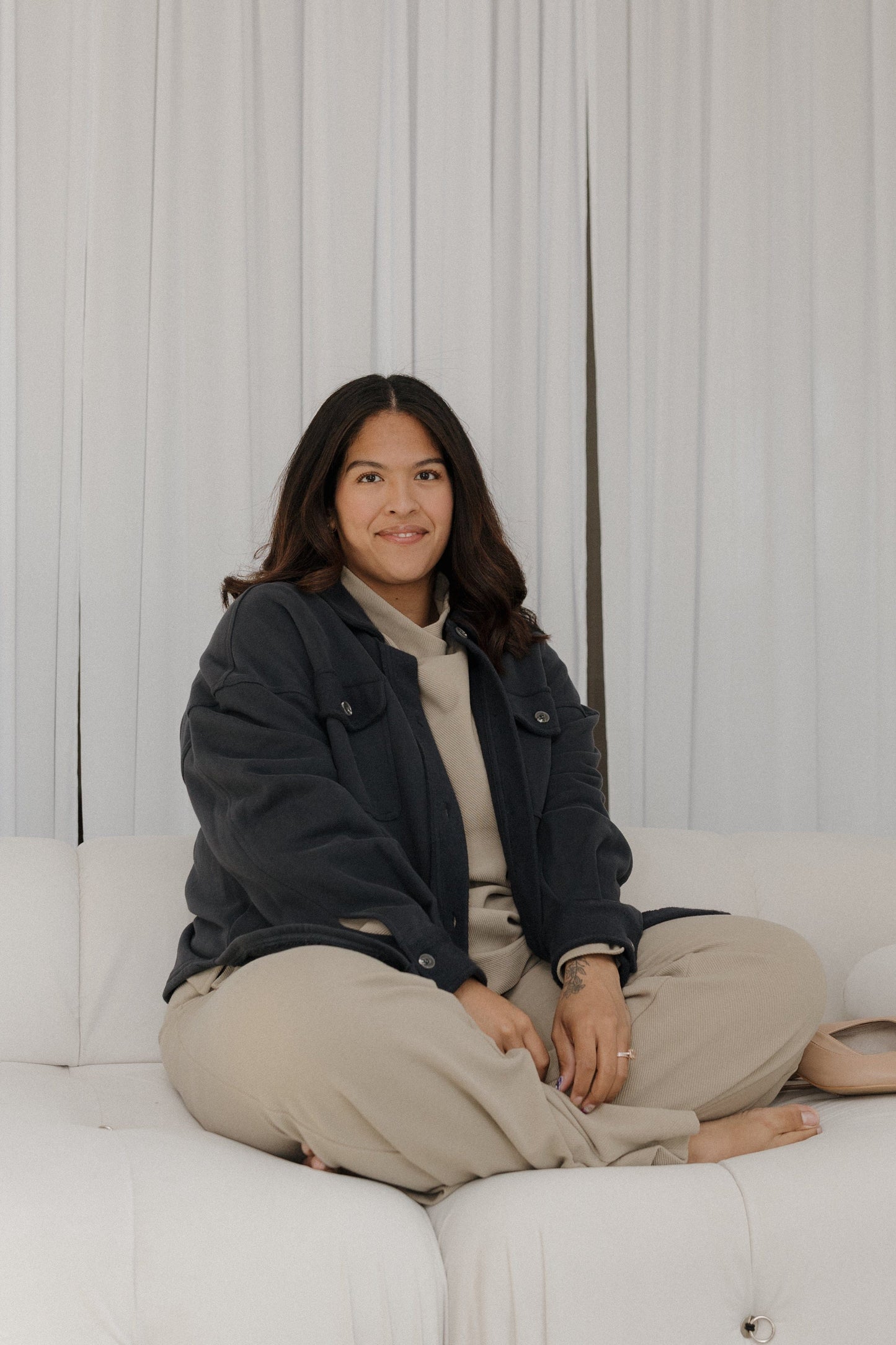 Person sitting on a white couch wearing a dark jacket and beige pants against a light gray background