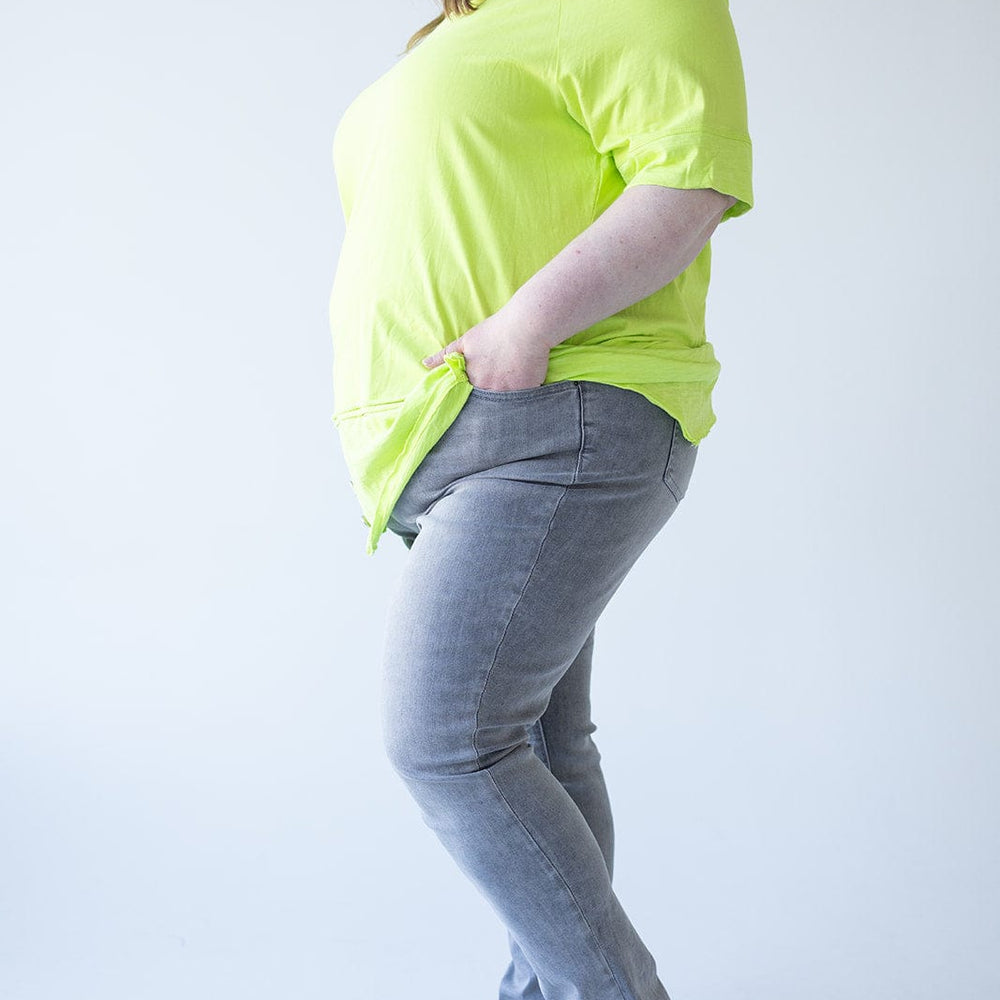 A person stands in profile wearing a bright yellow T-shirt, Love Marlow’s MID-RISE SKINNY JEAN WITH ZIPPER DETAIL IN LIGHT GREY, and black shoes, with their hands in their pockets.