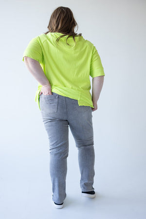 A person with long hair, wearing a bright green shirt, Love Marlow's MID-RISE SKINNY JEAN WITH ZIPPER DETAIL IN LIGHT GREY, and black shoes, stands facing away with one hand in a back pocket.