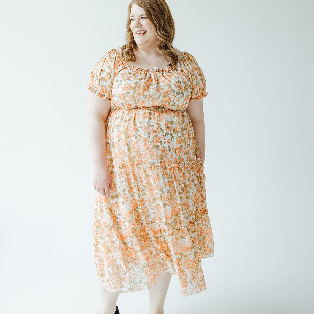 A woman stands smiling in a light-colored room, wearing the Mikarose MIDI-LENGTH FLORAL DRESS WITH BUBBLE SLEEVE and black shoes.