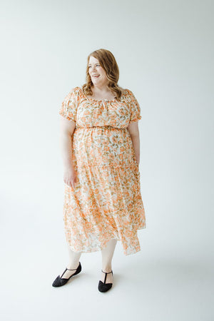 A woman stands smiling in a light-colored room, wearing the Mikarose MIDI-LENGTH FLORAL DRESS WITH BUBBLE SLEEVE and black shoes.