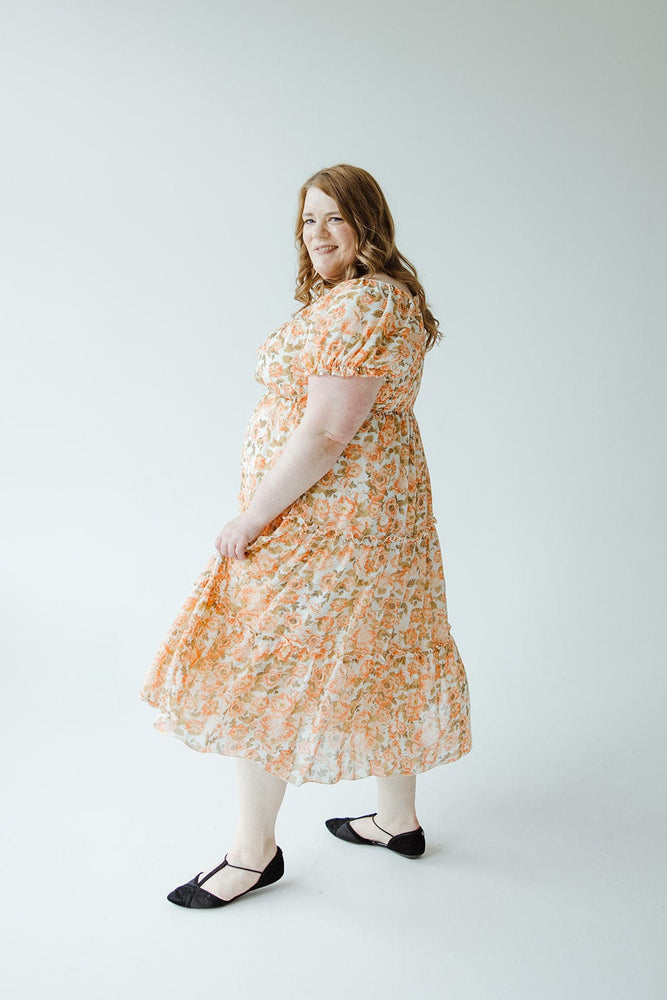 A woman in a Mikarose MIDI-LENGTH FLORAL DRESS WITH BUBBLE SLEEVE and black shoes stands in a bright, minimalist space, smiling and looking over her shoulder.