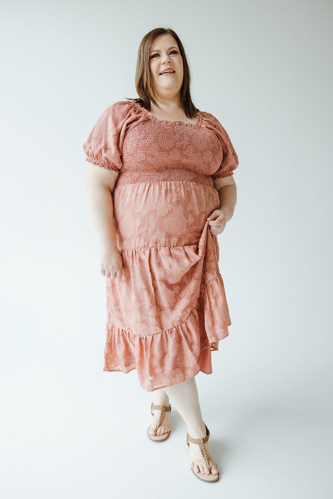 A woman in a MIDI-LENGTH LACE APPLIQUES IN PRESSED FLOWER by Mikarose stands against a white background, holding up a portion of her tiered skirt. She is wearing tan sandals and looking towards the camera with a neutral expression.