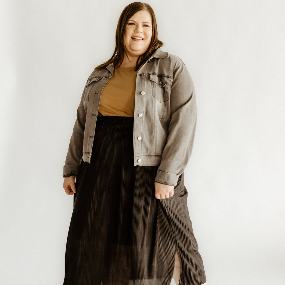 Person standing, smiling, wearing a gray jacket and tan top. She's styled in a black midi-length plissé skirt that shimmers subtly, paired with matching black shoes. Neutral background adds simplicity to her polished look.