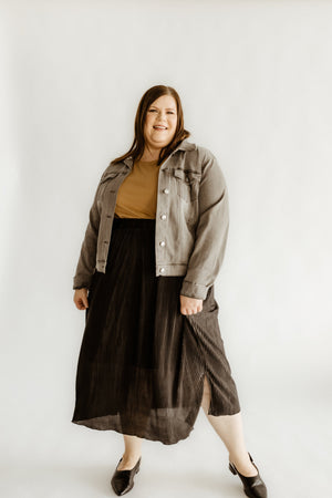 Person standing, smiling, wearing a gray jacket and tan top. She's styled in a black midi-length plissé skirt that shimmers subtly, paired with matching black shoes. Neutral background adds simplicity to her polished look.