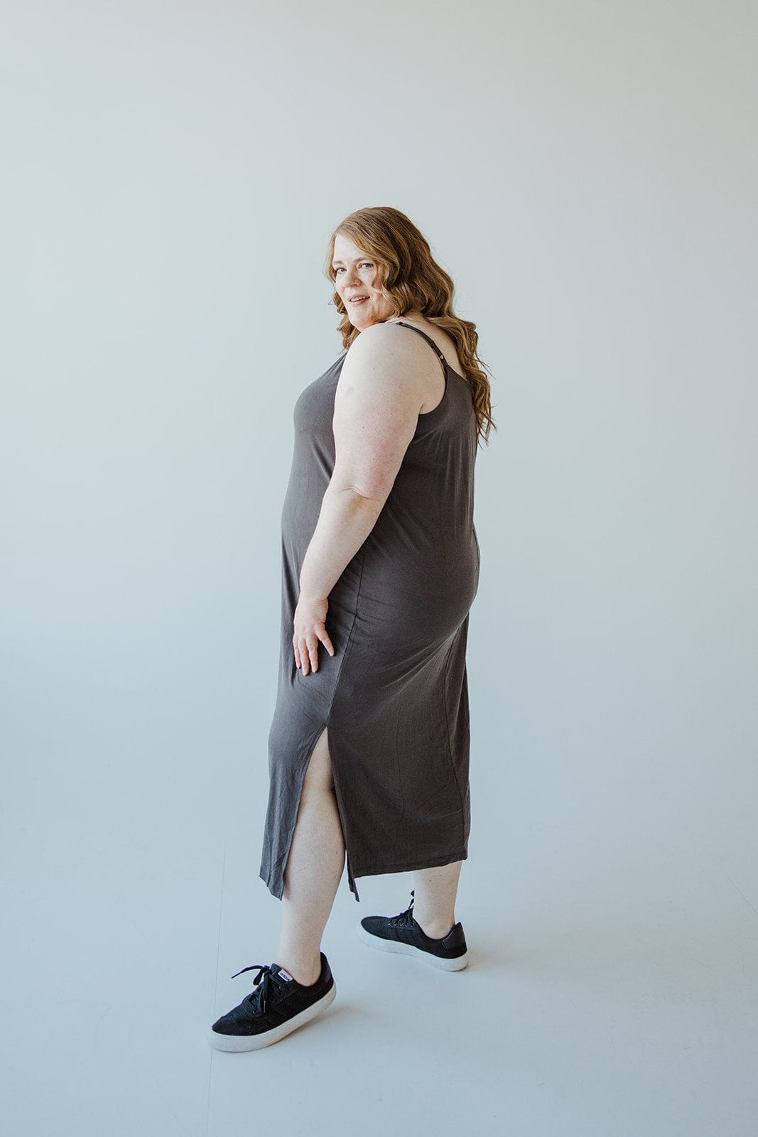 A woman in a dark modal fabric midi dress with adjustable cami straps and black sneakers poses sideways against a light background.