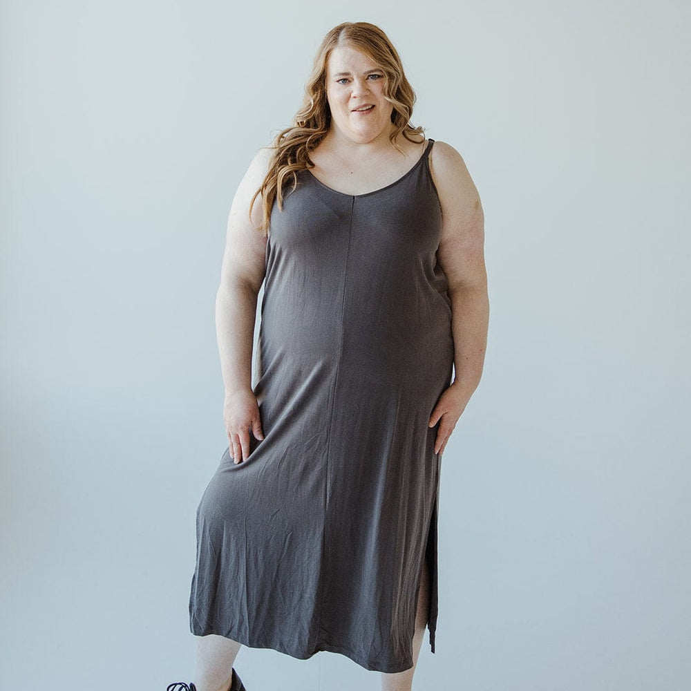 A person with flowing long hair is wearing a dark gray midi dress crafted from modal fabric with adjustable cami straps, paired with black sneakers, standing against a plain light background.