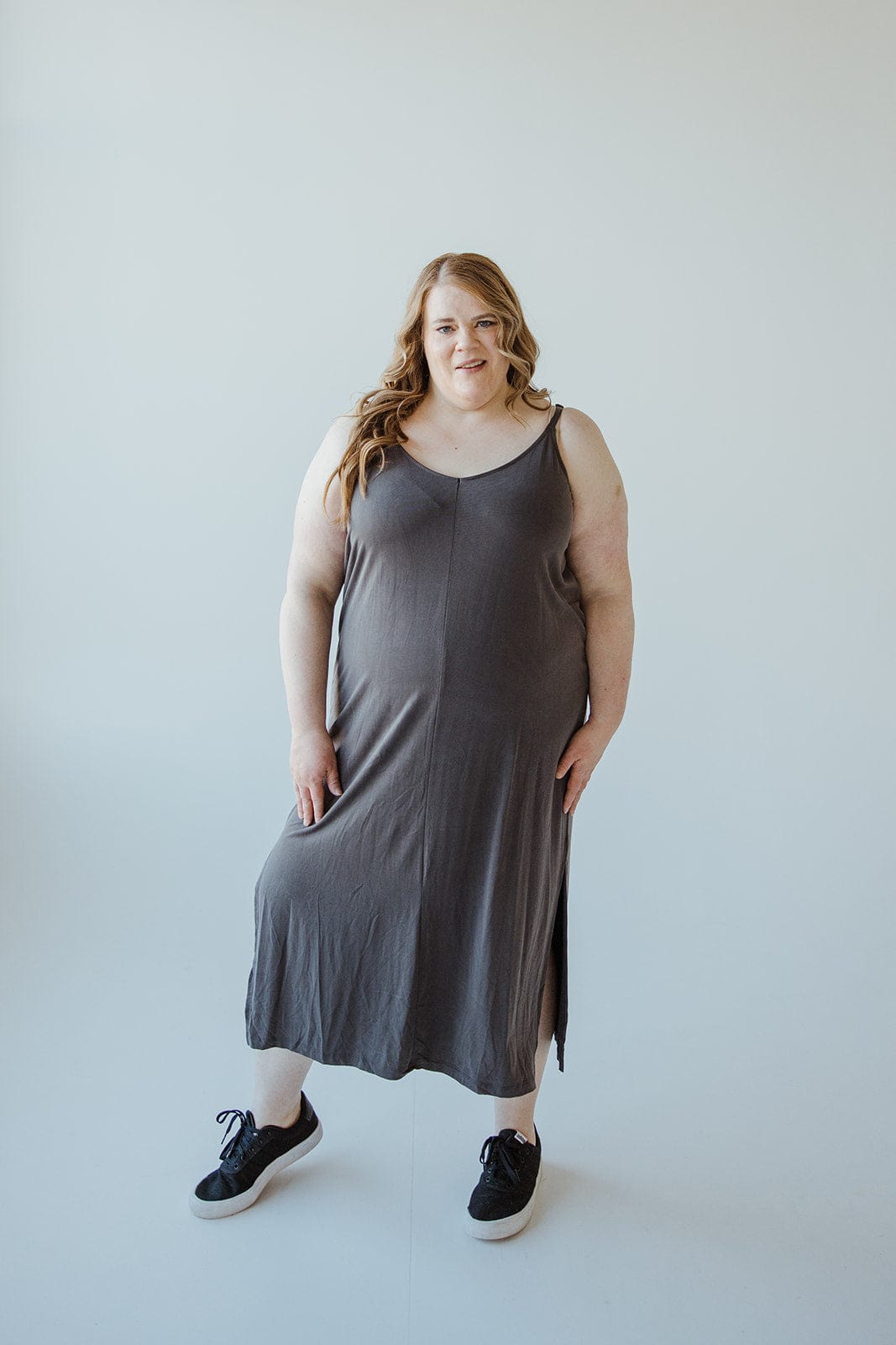 A person with flowing long hair is wearing a dark gray midi dress crafted from modal fabric with adjustable cami straps, paired with black sneakers, standing against a plain light background.