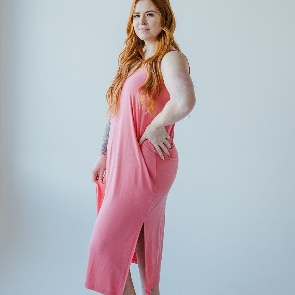 A person with long red hair wearing a sleeveless pink MIDI LENGTH SLIP DRESS IN CACTUS BLOSSOM by Love Marlow and black sandals stands against a plain white background.