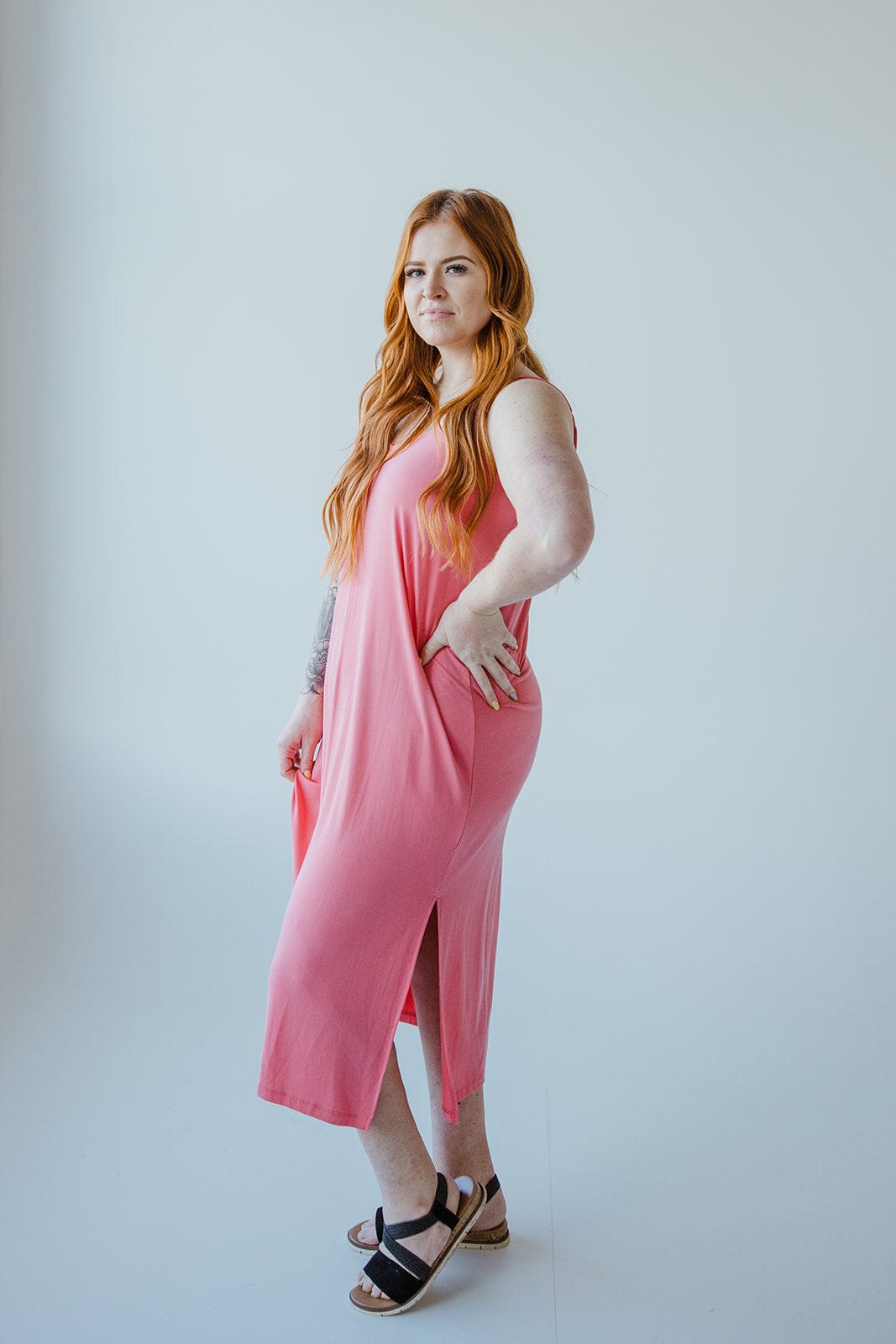 A person with long red hair wearing a sleeveless pink MIDI LENGTH SLIP DRESS IN CACTUS BLOSSOM by Love Marlow and black sandals stands against a plain white background.