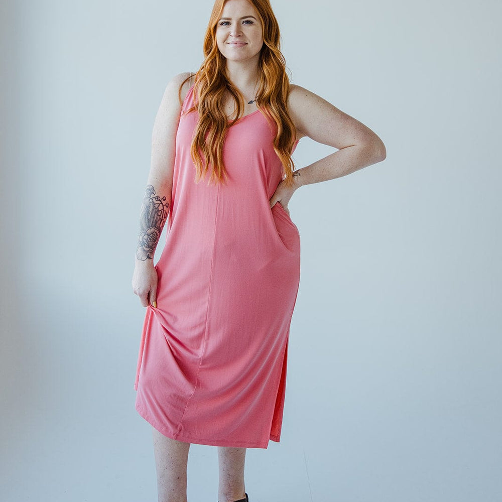 A woman with long red hair, wearing a sleeveless pink MIDI LENGTH SLIP DRESS IN CACTUS BLOSSOM from Love Marlow and black sandals, stands against a plain white background, smiling with one hand on her hip.