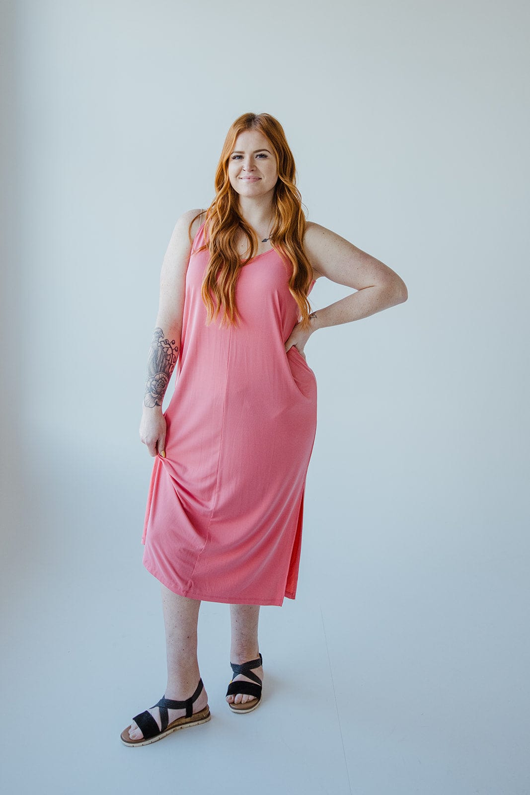 A woman with long red hair, wearing a sleeveless pink MIDI LENGTH SLIP DRESS IN CACTUS BLOSSOM from Love Marlow and black sandals, stands against a plain white background, smiling with one hand on her hip.