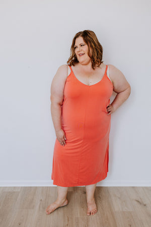 A smiling woman wearing a Love Marlow MIDI LENGTH SLIP DRESS IN SPICY CORAL with side slits stands barefoot on a wooden floor against a white wall.