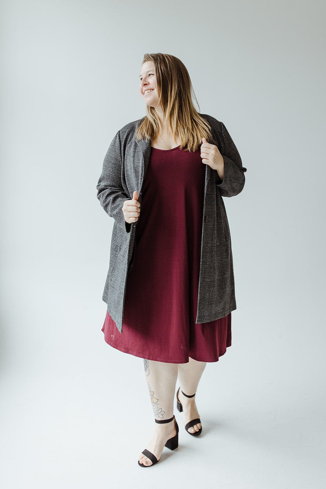 A person in a maroon dress and a plaid blazer stands confidently, smiling with one foot slightly forward on a plain background.