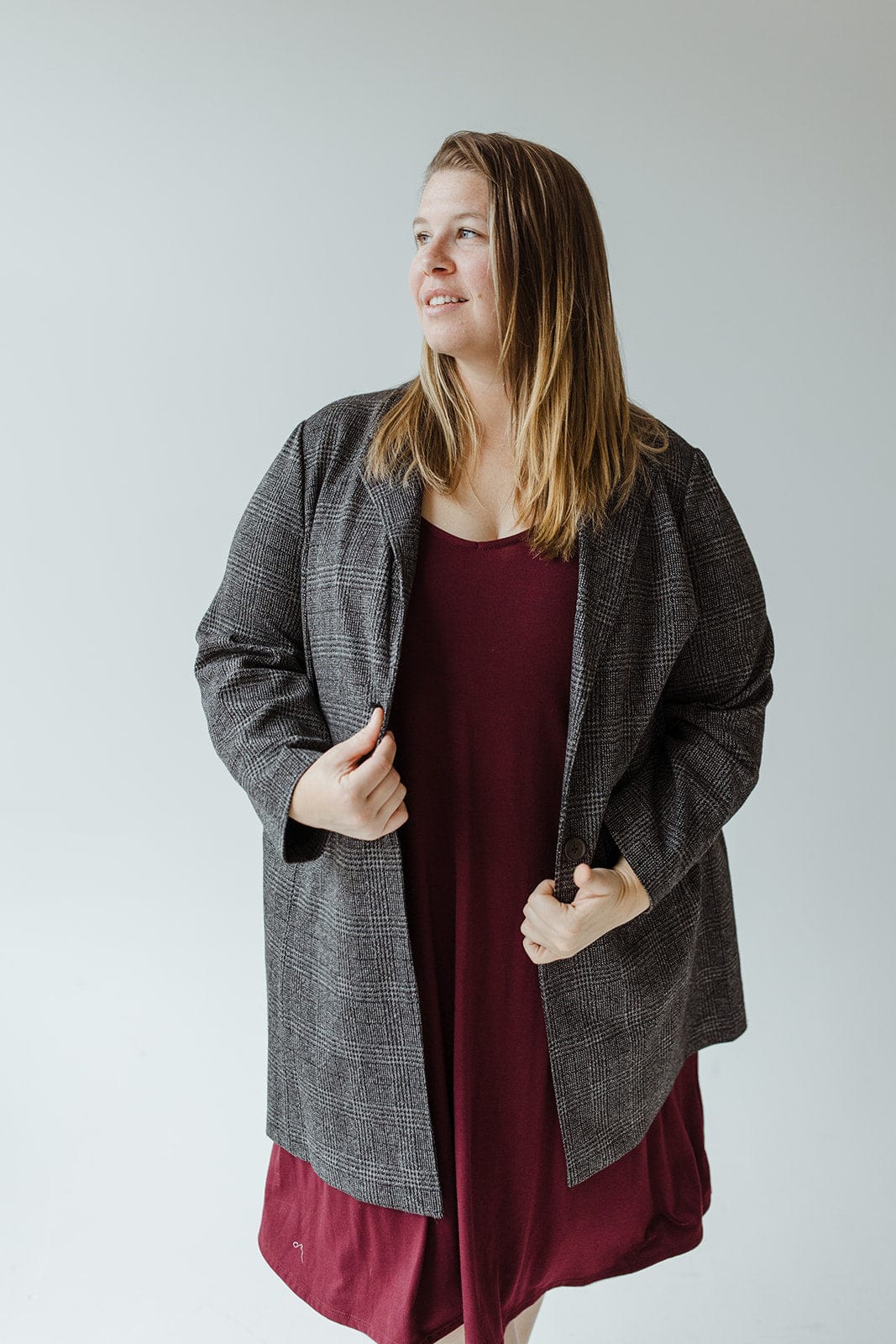 A person exudes elegance in a classic style, wearing a gray plaid blazer crafted from luxurious fabric over a burgundy dress, standing against a plain background.