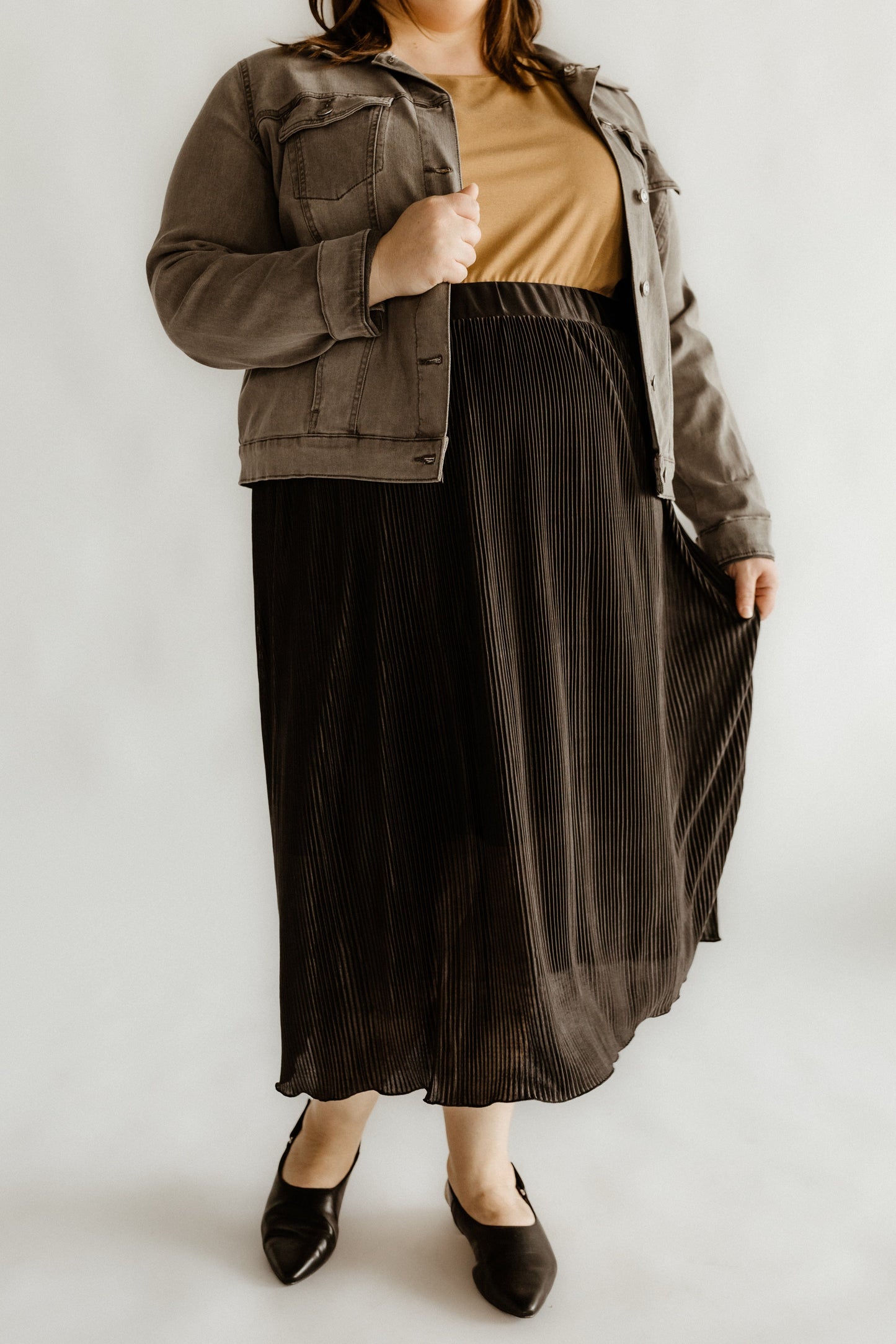 A woman wearing a gray jacket, beige top, and black pleated skirt of shiny plisse fabric stands against a plain background, gently holding the midi-length pleats with one hand.