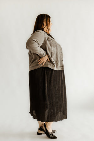 A person stands in profile, hands on hips, wearing a gray jacket over a sleek black dress. The dress features a MIDI LENGTH PLISSE SKIRT that shimmers gently with its shiny plisse fabric, adding an elegant touch to the ensemble.