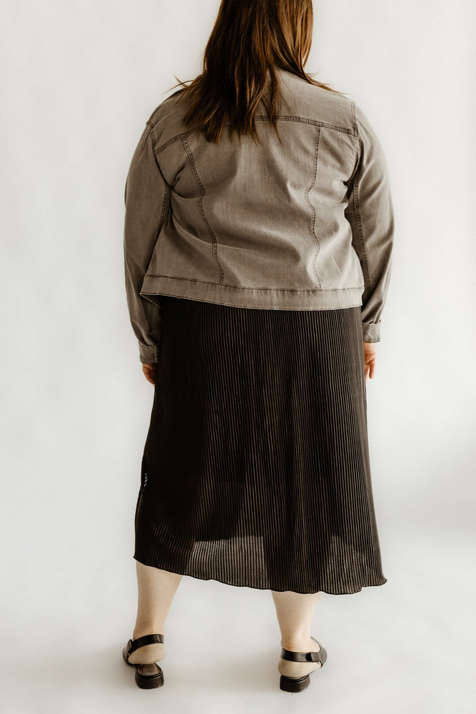 A person wearing a gray denim jacket, a black midi-length plissé skirt with an elastic waistband, and sandals is standing with their back to the camera.