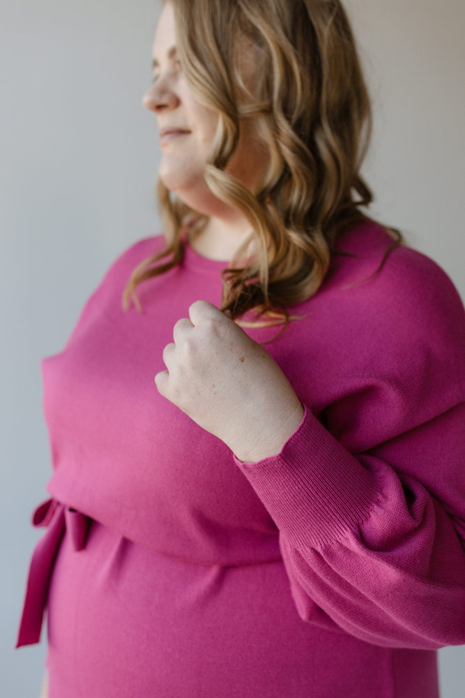 A woman in a fuchsia rose color MIDI LENGTH SWEATER DRESS by Yesta, with long wavy hair, stands in profile, her hand raised slightly. The vivid hue and bubble sleeves add a playful touch to her ensemble.