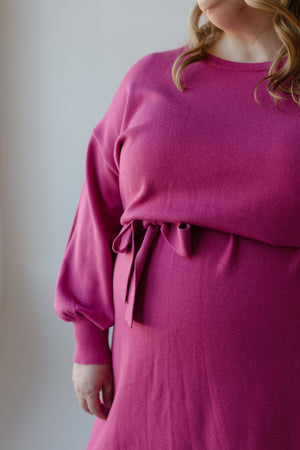 A person in a fuchsia rose MIDI LENGTH SWEATER DRESS, designed by Yesta, featuring long bubble sleeves and a ribbon tie detail.