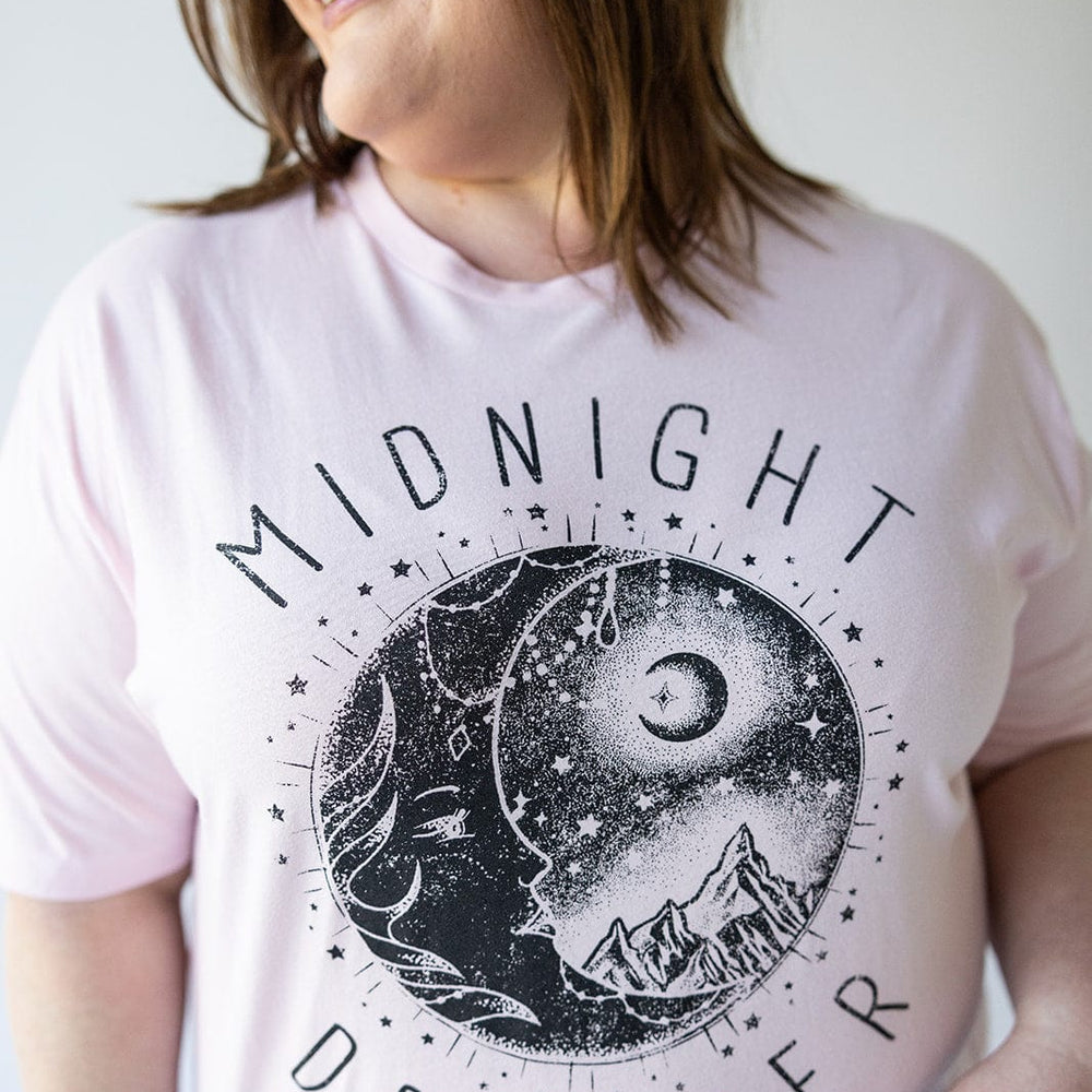 A person is wearing the "MIDNIGHT DREAMER" graphic tee in light pink by Love Marlow, featuring a moon, stars, and mountains design.