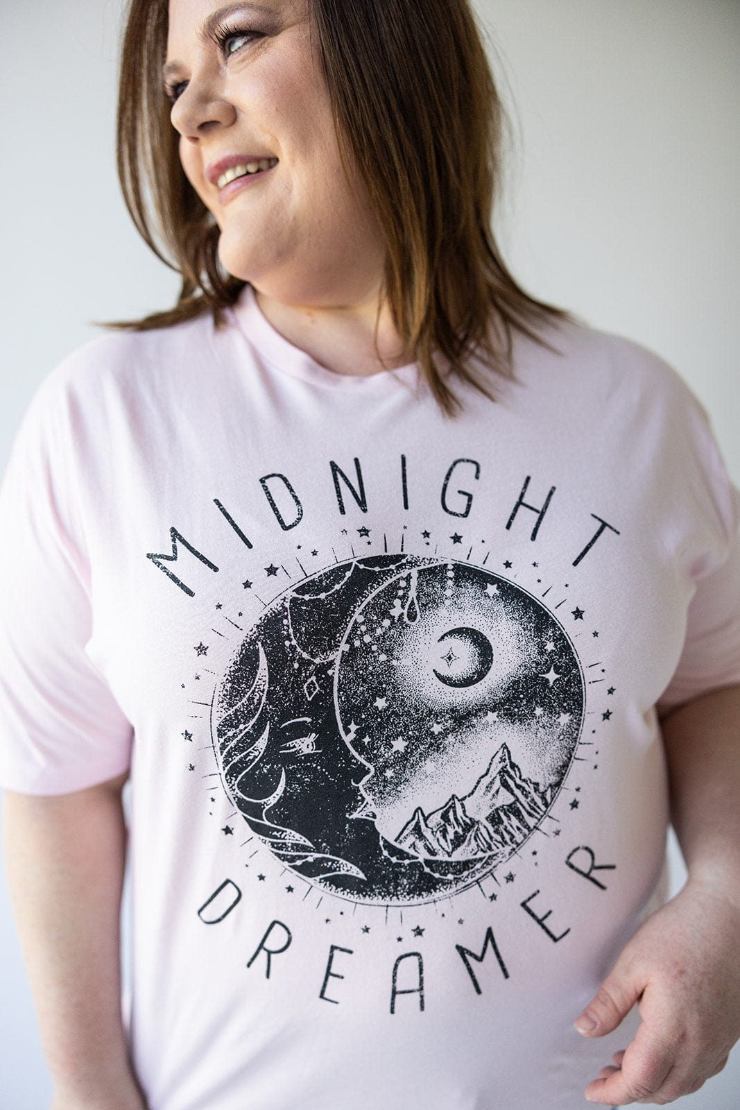 A person is wearing the "MIDNIGHT DREAMER" graphic tee in light pink by Love Marlow, featuring a moon, stars, and mountains design.