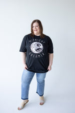 A person with brown hair is seen from the back, wearing Love Marlow's "MIDNIGHT DREAMER" graphic tee in black paired with blue jeans, standing against a plain white background.