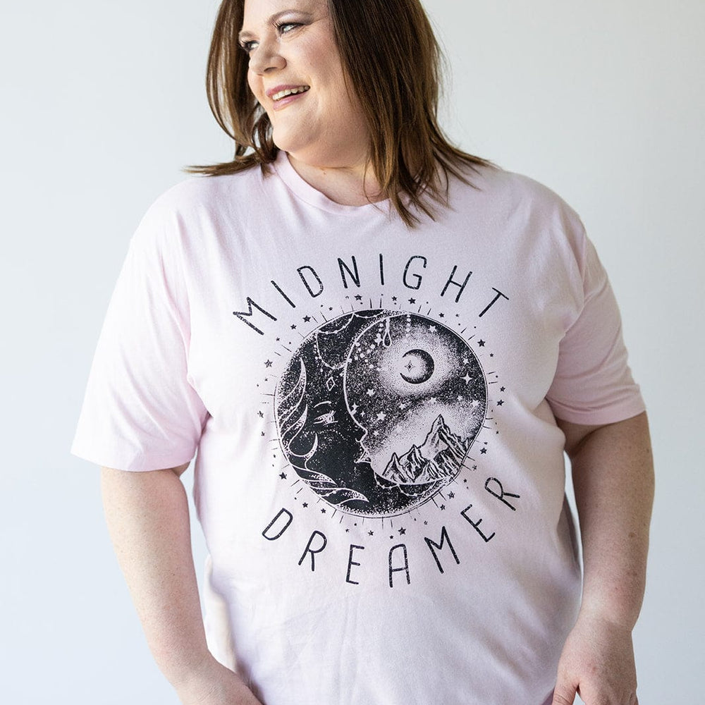 A person stands against a plain background, wearing Love Marlow's light pink "MIDNIGHT DREAMER" graphic tee, made from 100% cotton and featuring a serene design of the moon and mountains.