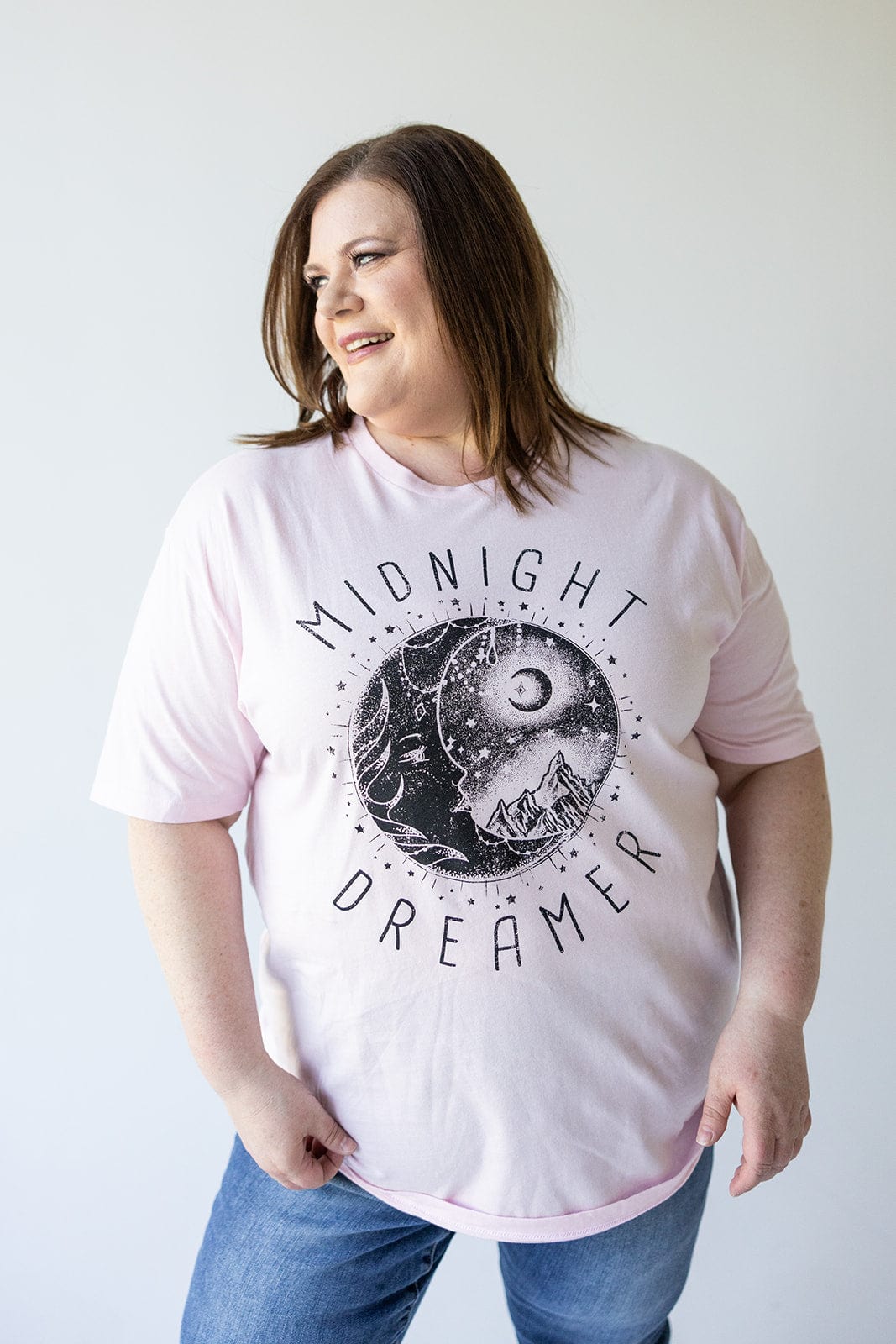 A person stands against a plain background, wearing Love Marlow's light pink "MIDNIGHT DREAMER" graphic tee, made from 100% cotton and featuring a serene design of the moon and mountains.