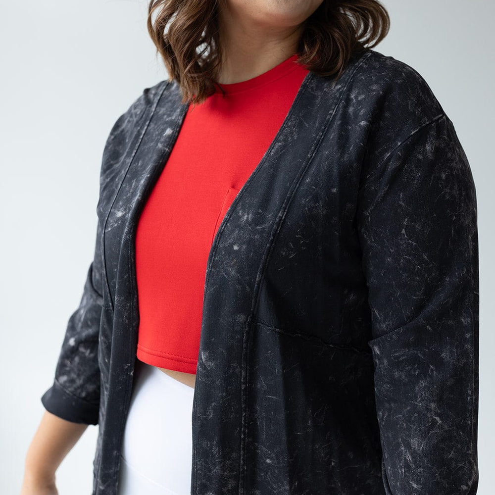 A person wearing a red top, black mid-hip length Mineral Wash Cardigan, and white pants smiles while looking to the side against a neutral background.