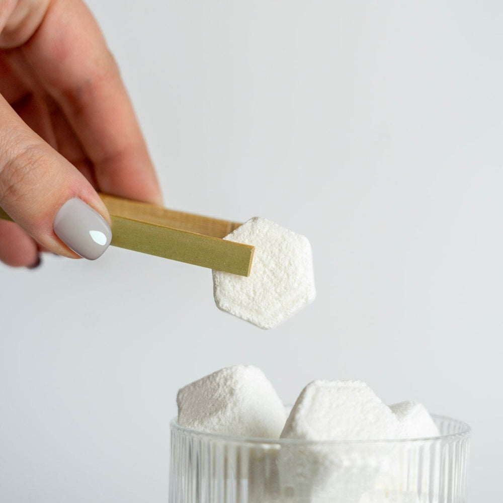 Hand holding a gold spoon over a glass filled with shower steamers on a light gray background