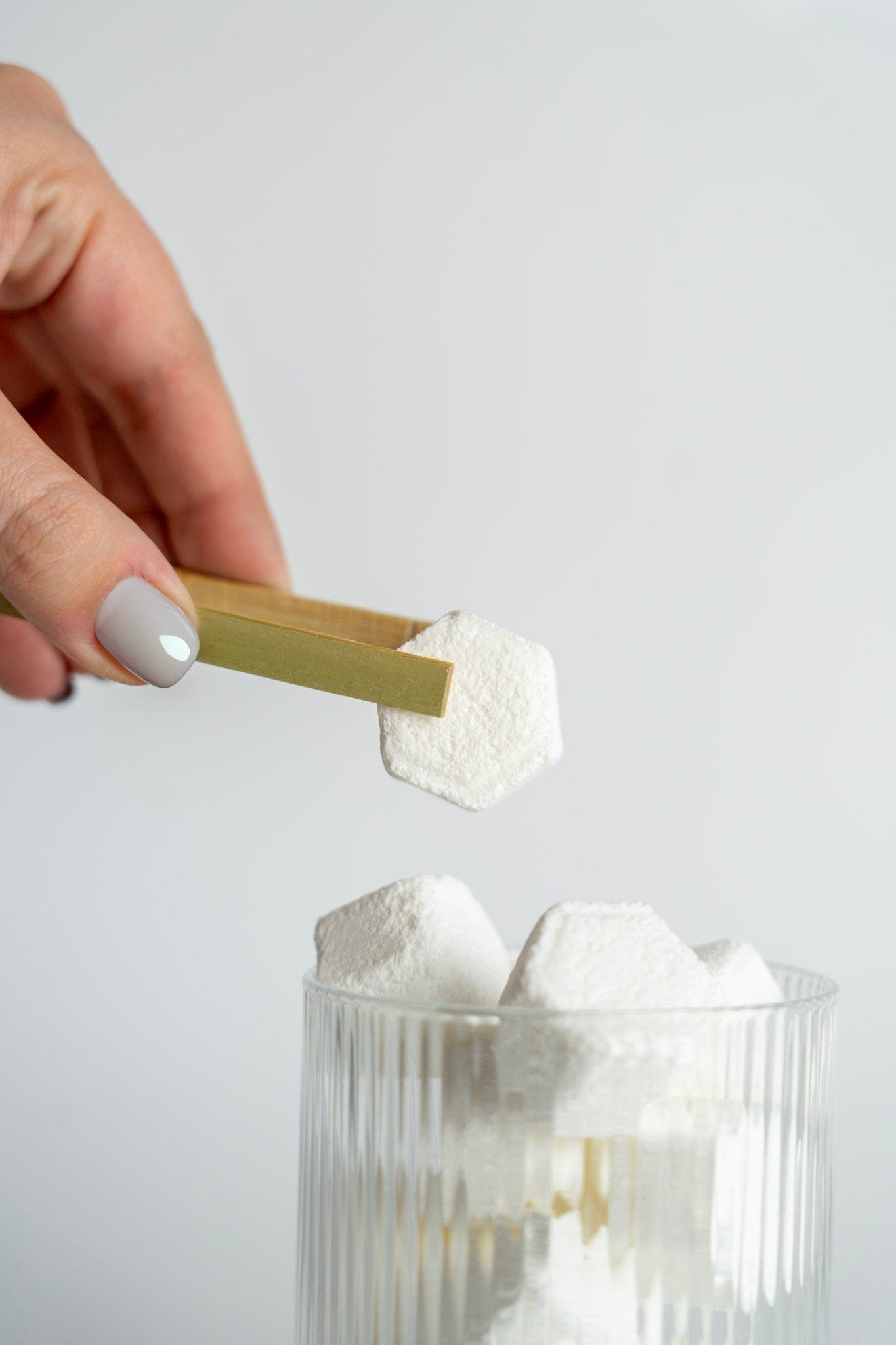 Hand holding a gold spoon over a glass filled with shower steamers on a light gray background
