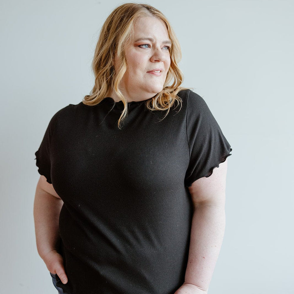 A person with light hair wearing a stylish black shirt and jeans, with a hint of sophistication from its mock neck ribbed design, stands against a plain background.