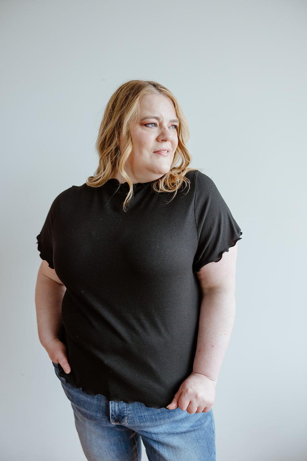 A person with light hair wearing a stylish black shirt and jeans, with a hint of sophistication from its mock neck ribbed design, stands against a plain background.