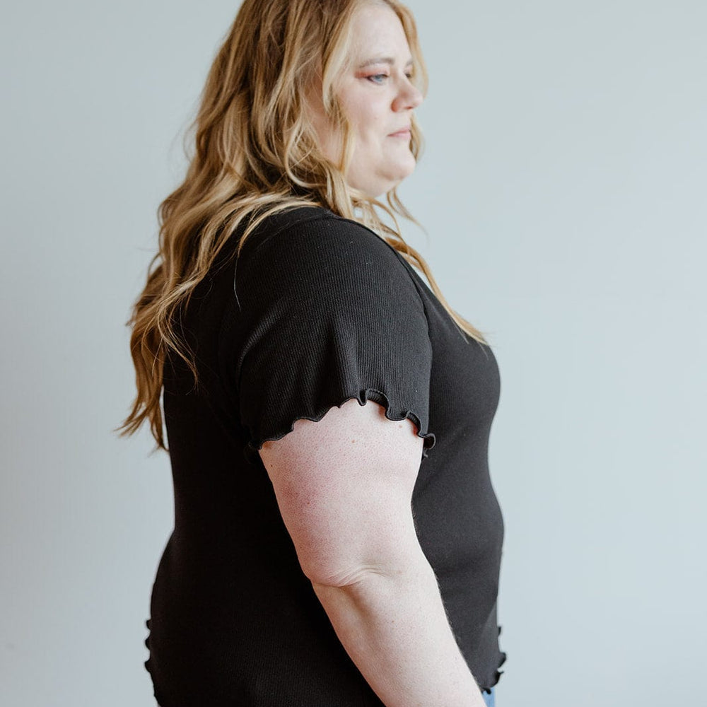 Person with long hair wearing a stylish black shirt and jeans, seen from the side against a plain background.