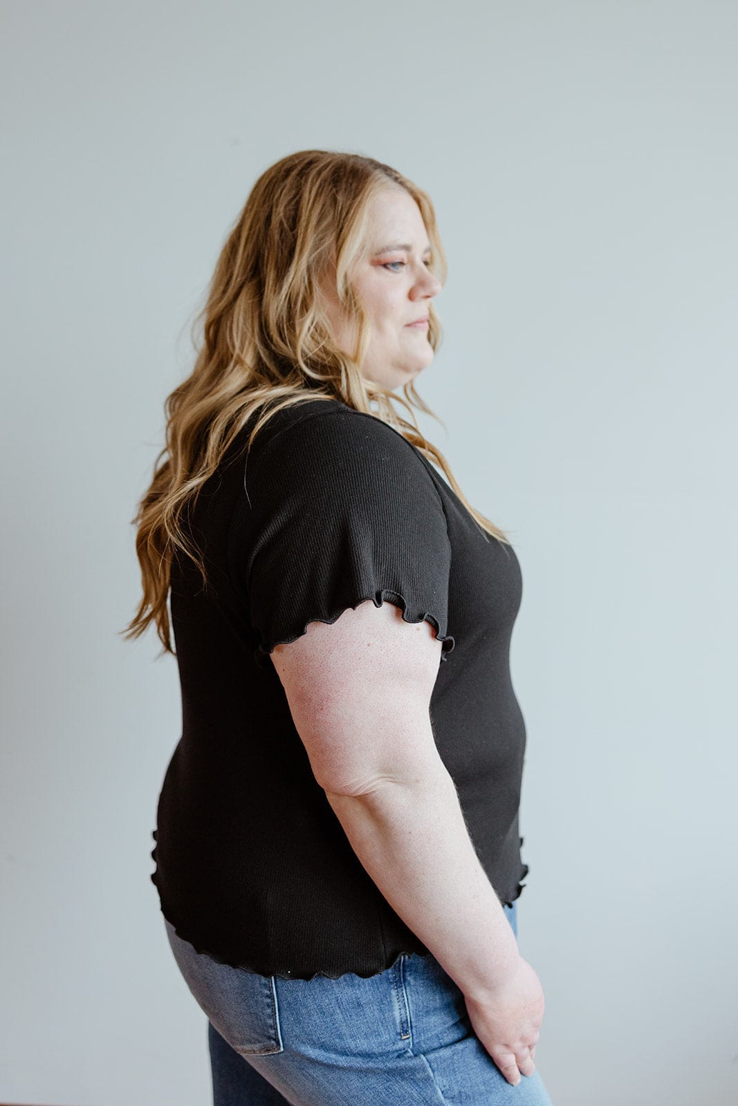 Person with long hair wearing a stylish black shirt and jeans, seen from the side against a plain background.