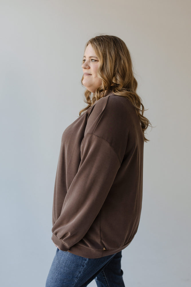 A person with long hair, dressed in a brown Yesta MOCK TURTLENECK ZIPPER DETAIL BLOUSE and jeans, stands sideways against a plain background.