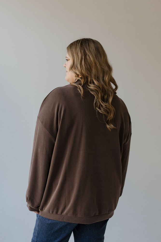 A person with long wavy hair is wearing a brown Yesta mock turtleneck zipper detail blouse and jeans, facing away against a neutral background.