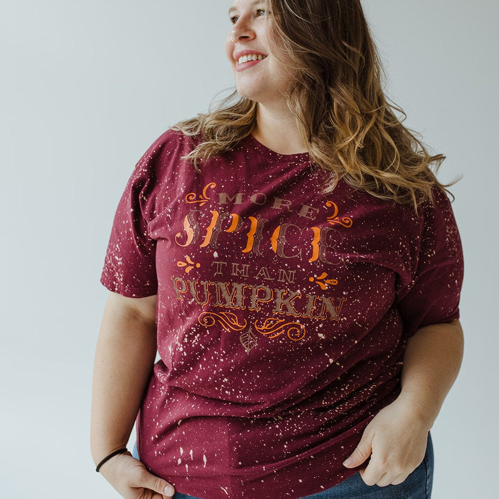 A person confidently stands against a plain background, wearing Southern Bliss's "MORE SPICE THAN PUMPKIN" graphic tee in maroon, featuring a pumpkin spice theme. This tee is beautifully hand bleached for a unique look and paired with blue jeans. It's an ideal style choice for mid to plus-size fashion enthusiasts who adore autumn vibes.