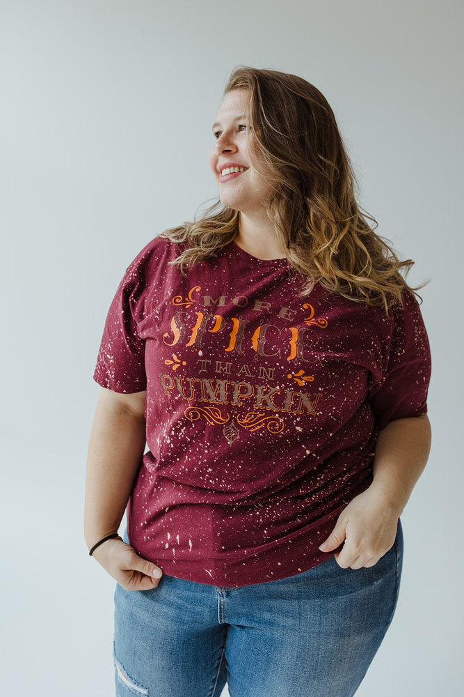 A person confidently stands against a plain background, wearing Southern Bliss's "MORE SPICE THAN PUMPKIN" graphic tee in maroon, featuring a pumpkin spice theme. This tee is beautifully hand bleached for a unique look and paired with blue jeans. It's an ideal style choice for mid to plus-size fashion enthusiasts who adore autumn vibes.