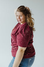 A mid to plus-size person with long hair is facing away, wearing the Southern Bliss "MORE SPICE THAN PUMPKIN" graphic tee in hand-bleached maroon speckled design, paired with blue jeans.