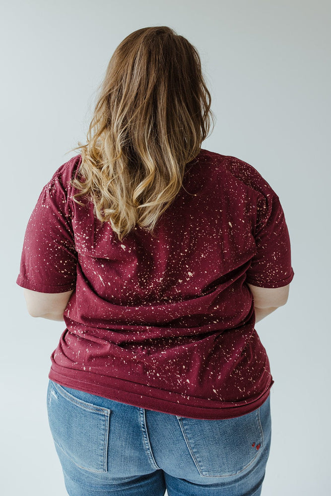 A mid to plus-size person with long hair is facing away, wearing the Southern Bliss "MORE SPICE THAN PUMPKIN" graphic tee in hand-bleached maroon speckled design, paired with blue jeans.