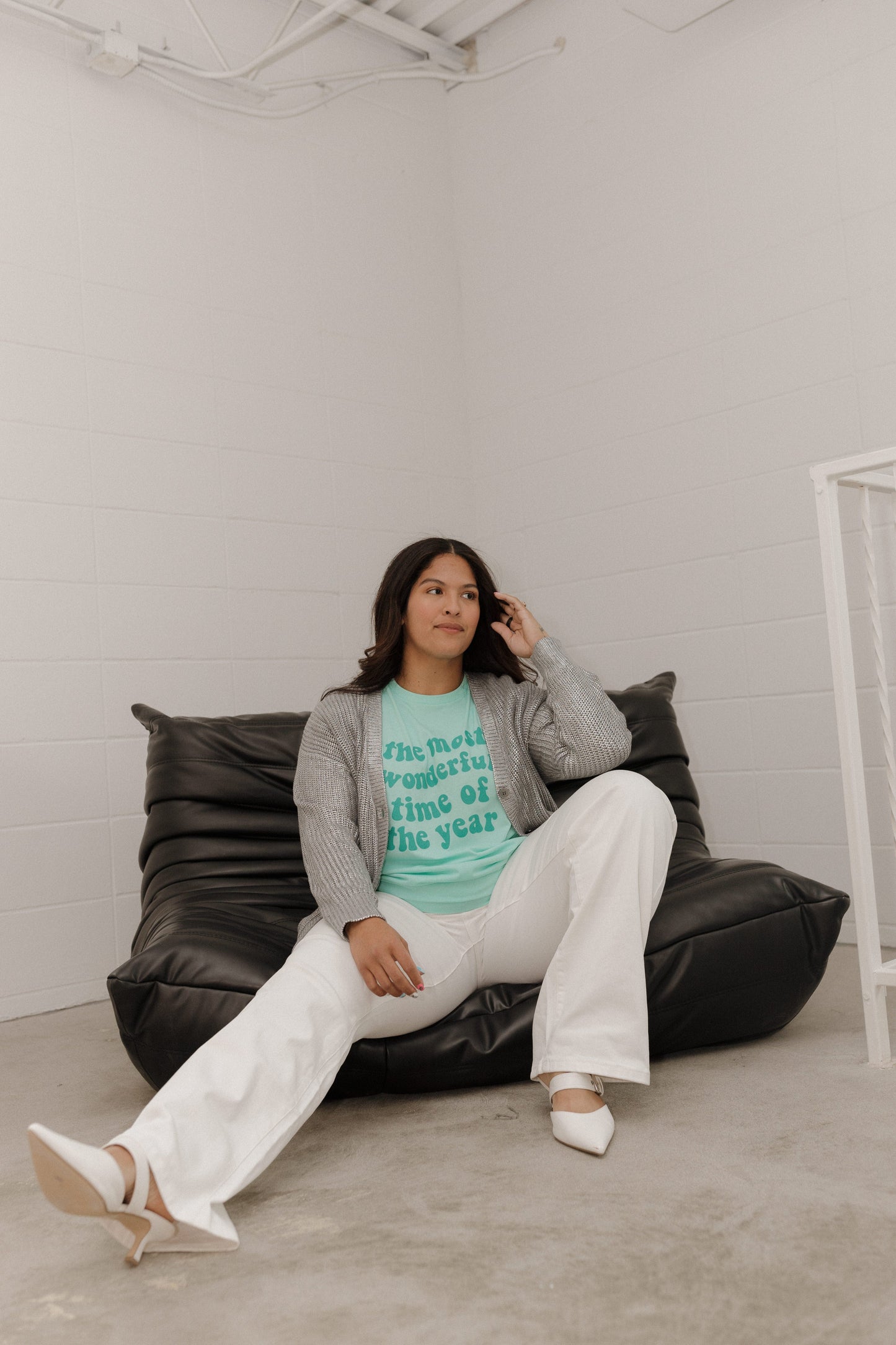 Woman sitting on a black bean bag chair wearing a gray sweater and white pants in a minimalistic room.