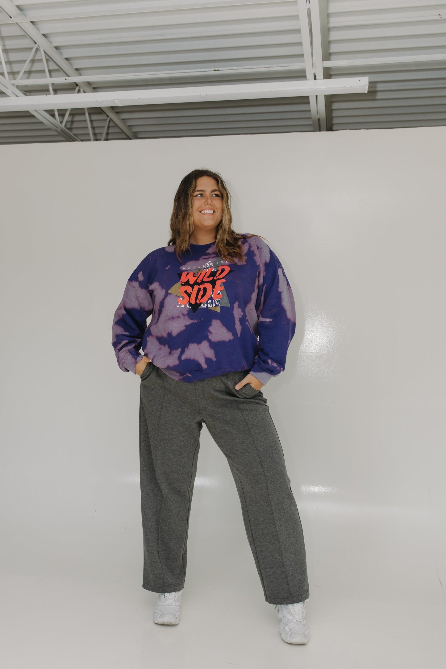 Person wearing a tie-dye sweatshirt with colorful text and gray pants in an indoor setting.
