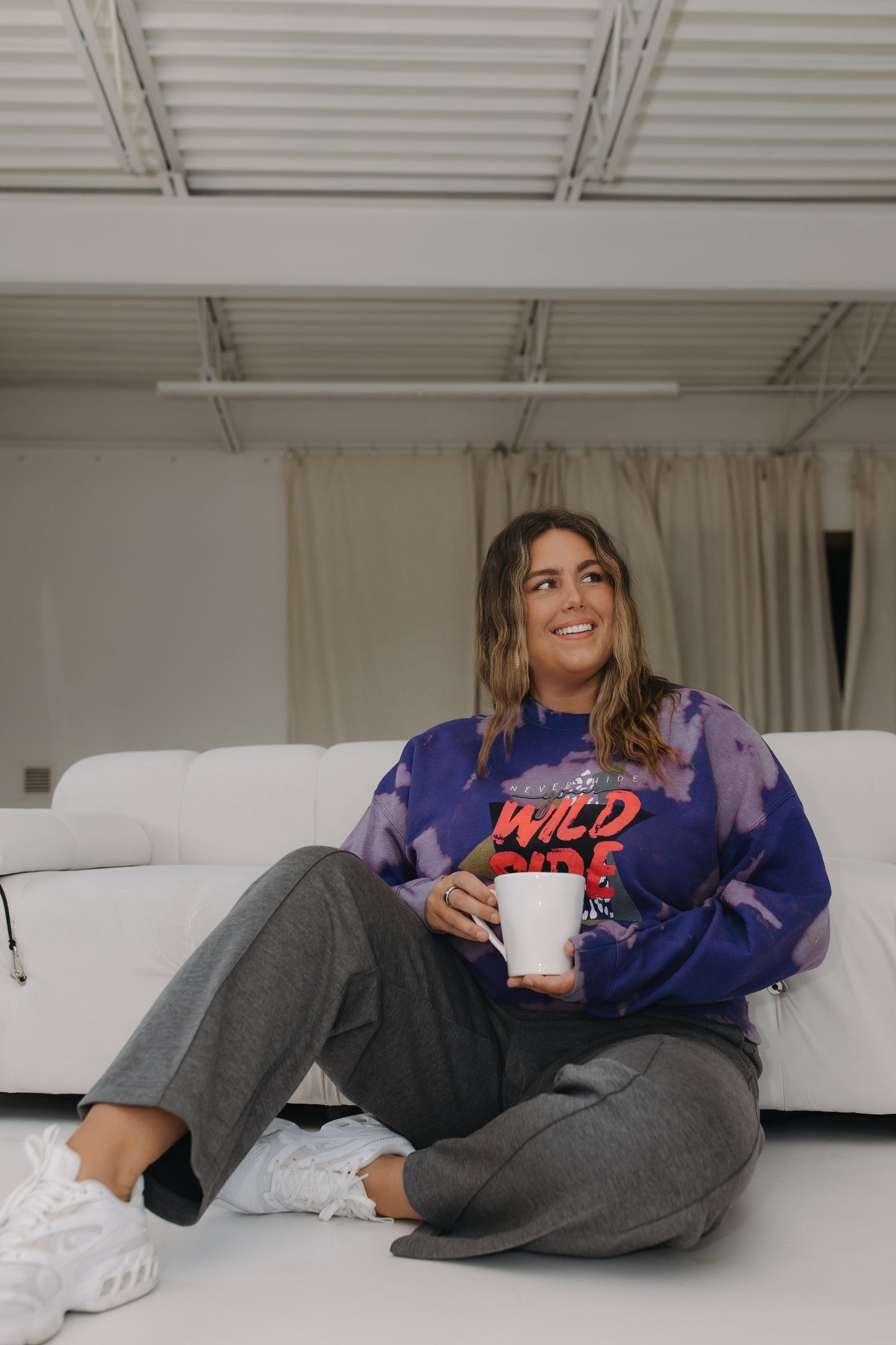 Woman sitting on a white couch holding a white mug, wearing a colorful sweatshirt and gray pants.