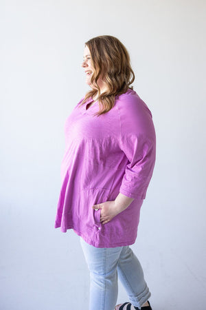 A person with long hair wearing a luxurious cotton, loose-fitting pink top (NOTCHED NECK TUNIC IN VERBENA by Love Marlow) and light-colored jeans stands facing left, smiling.