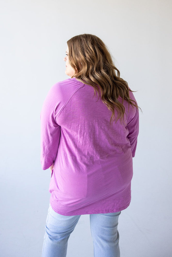 A woman with wavy hair, wearing a luxurious Love Marlow NOTCHED NECK TUNIC IN VERBENA and light blue jeans, is seen from the back against a plain background.