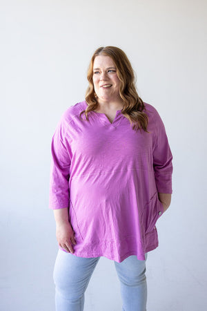 A person wearing a bright pink NOTCHED NECK TUNIC IN VERBENA by Love Marlow and light blue jeans stands against a plain white background, looking slightly to the side and smiling.