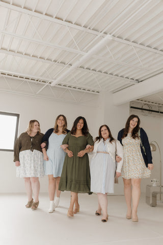 Five women standing in a row wearing various dresses in a bright room with white ceiling.